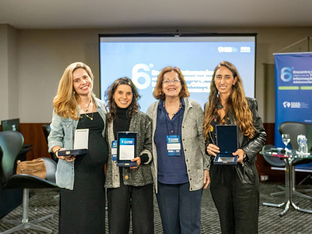 Quatro mulheres posam lado a lado, cada uma com uma placa comemorativa nas mãos, diante do telão do 6º Encontro Internacional.