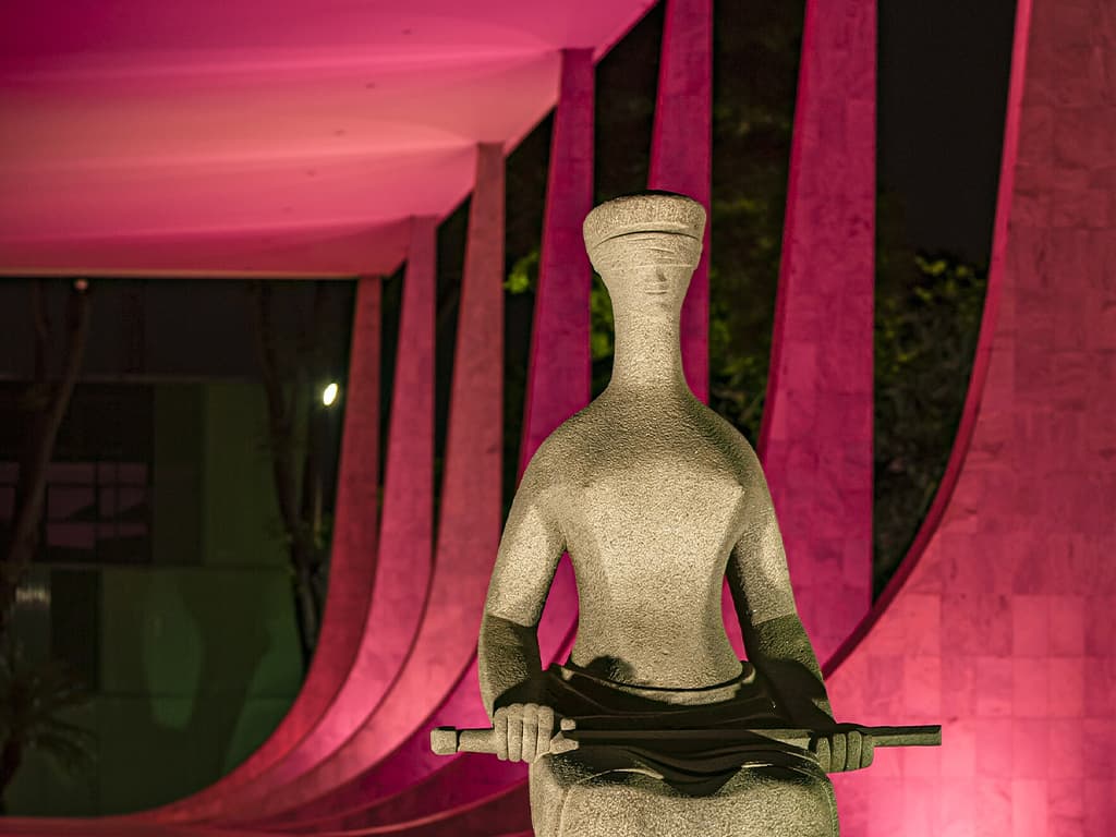 A estátua “A Justiça”, de Alfredo Ceschiatti, diante do Supremo Tribunal Federal, com os arcos do edifício iluminados em rosa à noite.