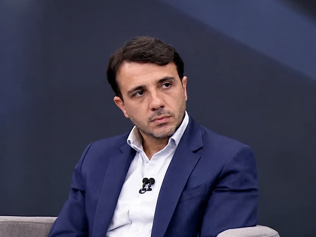 Renan Santos, de terno azul e camisa branca com microfone de lapela, sentado durante uma entrevista.