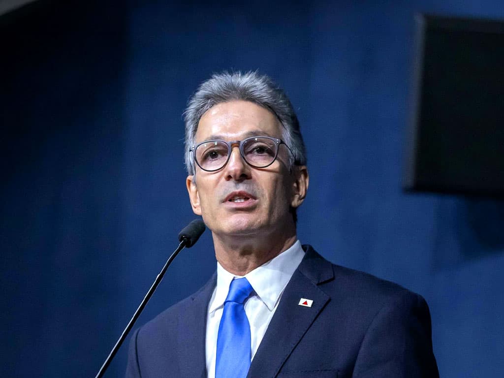 Romeu Zema, de terno escuro, camisa branca e gravata azul, fala ao microfone diante de um fundo azul.
