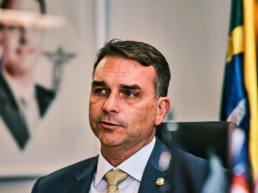 Flávio Bolsonaro, de terno escuro, camisa clara e gravata dourada, sentado em um gabinete com a bandeira do Brasil ao fundo.